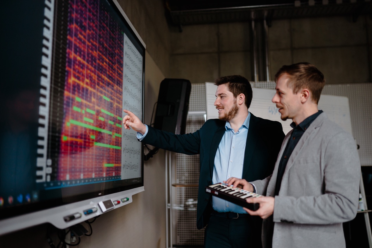 Sebastian Murgul und Alexander Lüngen demonstrieren am Klangio Transcription Studio, wie die KI komplexe Mehrspur-Aufnahmen analysiert und in Notation überführt. Die farbcodierte Darstellung zeigt erkannte Tonhöhen, Timing-Informationen und Instrumentenzuweisungen in Echtzeit.