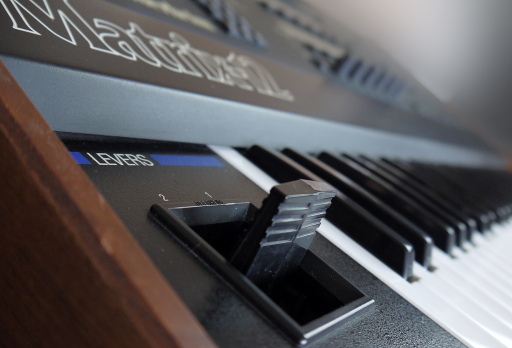 Nahaufnahme eines Oberheim Matrix-12 Synthesizers mit fokussiertem Lever-Hebel links neben der Tastatur und unscharfem Matrix-12-Schriftzug im Hintergrund.