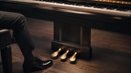Drei goldene Piano-Pedale unter einem schwarzen Klavier, daneben der Fuß eines Pianisten.