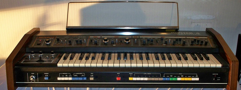 Roland Jupiter-4 Vintage-Synthesizer mit Holzseitenteilen, Tastatur, Bedienfeld und farbigen Preset-Tastern in Gesamtansicht.