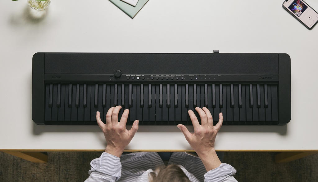 Casio CT-FS1BK mit vollständig schwarzer Tastatur beim beidhändigen Spielen, von oben auf einem weißen Tisch fotografiert.